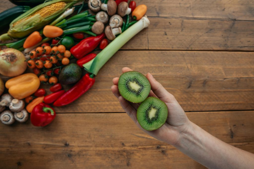 A hand holds sliced kiwi above a wooden table with fresh vegetables, highlighting healthy ingredients.