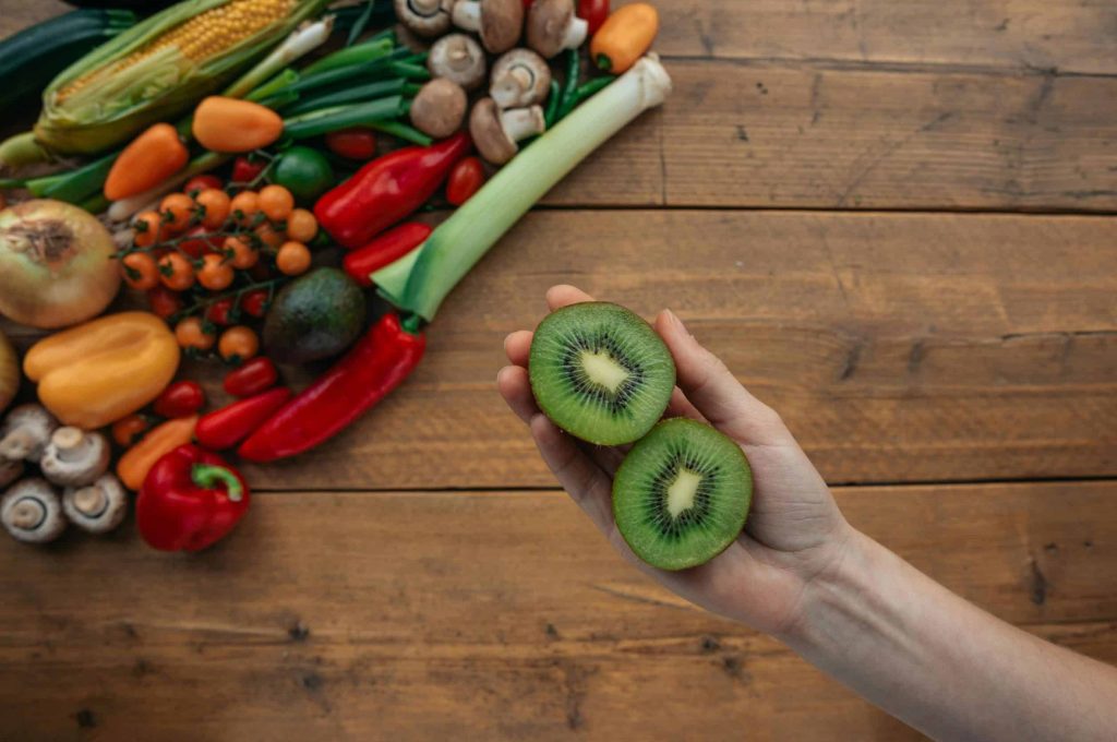 A hand holds sliced kiwi above a wooden table with fresh vegetables, highlighting healthy ingredients.